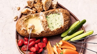 Grote broodbol gevuld met twee soorten kaasfondue, geserveerd met stukjes brood, cherrytomaten, komkommer en oranje paprika op een houten schaal