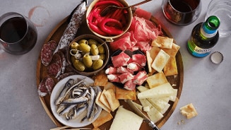 Tapas plank met diverse hapjes zoals gedroogde worsten, olijven, sardines, gesneden kaas, crackers, en serranoham, vergezeld van een glas rode wijn en een flesje bier.