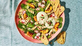 Salade met gegrilde witte asperges, gegrilde perzik, radijs, spinazie, burrata en krokante toast op een roze bord.