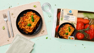 Lasagne op een zwart bord, naast een AH lasagnepakket met verse groente, verpakking en een glas water op een lichtgroene ondergrond.