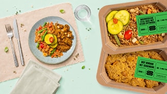 Bord met Indonesische maaltijd van rijst, groenten, vlees en augurk, naast twee kartonnen verpakkingen met labels "Indonesische Stijl Nasi" en "Beef Rendang" van Albert Heijn.