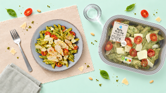 Pasta pesto kip maaltijdsalade met stukjes kip, cherrytomaten, kaas, pijnboompitten en basilicum, geserveerd op een bord en in de originele Albert Heijn verpakking.