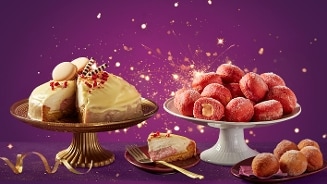 Feestelijke taart met witte chocoladeglazuur en decoratie op een gouden schaal naast een schaal met roze gevulde mini-donuts tegen een paarse achtergrond met fonkelende lichtjes