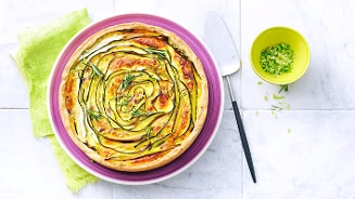 Groententaart met courgette en kruiden, geserveerd op een paarse schaal met een groene servet en een bakje gesneden groene kruiden op de achtergrond.