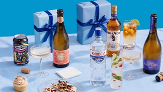 Verschillende alcoholvrije dranken, waaronder vandeStreek Non Alcoholic IPA, Vintense, Gordon’s 0.0% gin en Captain Morgan 0.0% rum, geserveerd in glazen, naast hapjes, cupcakes en twee blauw ingepakte cadeaus op een blauwe ondergrond.