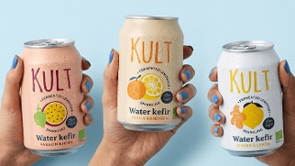 Drie handen houden blikjes Kult Water Kefir vast in de smaken Passion & Hops, Yuzu & Mandarin en Ginger & Lemon, tegen een lichtblauwe achtergrond.