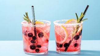 Twee glazen roze cocktail met ijs, citroen, blauwe bessen, rozemarijn en metalen rietjes, tegen een blauwe achtergrond.