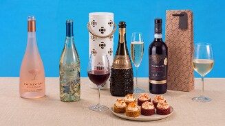 Verschillende flessen wijn, een fles mousserende wijn, wijnglazen en een schaal met luxe hapjes op een tafel met blauwe achtergrond.