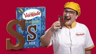 Verkade melk chocoladeletter S in blauwe verpakking met een persoon in witte Verkade outfit en gele baret tegen een rode achtergrond