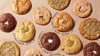 Verschillende soorten grote koekjes, waaronder loaded cookie pistache, loaded cookie s'mores, cookie hazelnoot witte chocolade en koeken met roze glazuur, liggen op bruin papier.