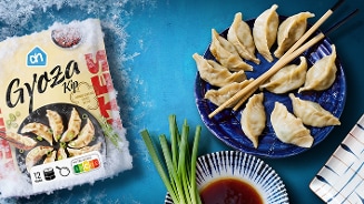 AH Gyoza Kip diepvriesverpakking naast een blauw bord met kip gyoza dumplings, sojasaus en bosui op een blauwe ondergrond