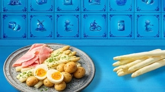 Bord met witte asperges, gekookte aardappelen, half gesneden gekookte eieren en plakjes ham, op een blauwe ondergrond met decoratieve tegeltjes op de achtergrond. Er liggen daarnaast losse witte asperges.