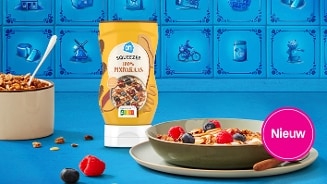 Een AH Squeezer 100% pindakaas staat centraal op een tafel, omringd door een kom ontbijtgranen en een bord met yoghurt, granola, pindakaas en vers fruit zoals frambozen en blauwe bessen. Op de voorgrond staat een roze label met de tekst "Nieuw". De achtergrond bestaat uit blauwe tegels met Hollandse motieven.