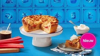 Alt tekst: Rabarberkruimeltaart op een witte taartstandaard, met een stuk taart geserveerd op een bordje naast een schaaltje slagroom. Op de achtergrond een blauw tegelpatroon en links verse rabarberstelen en een glas koffie. Cirkel met "Nieuw" benadrukt de introductie van het product.