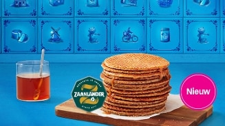 Stapel Zaanlander stroopwafels op een houten plank naast een glas thee, met blauwe Hollandse tegeltjes op de achtergrond en een roze label met "Nieuw".