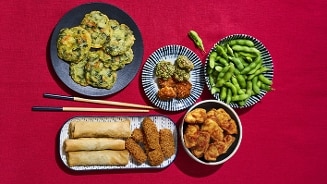 Assortiment van Aziatische voorgerechten op een rode achtergrond, met onder andere edamame, loempia's, tempura en gevarieerde hapjes.