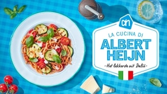 Bord met Italiaanse spaghetti met courgette, tomaat en basilicum naast een bordje met Parmezaanse kaas en een bord met het logo van Albert Heijn en de tekst "La cucina di Albert Heijn - Het lekkerste uit Italië" op een blauw geruit tafelkleed.