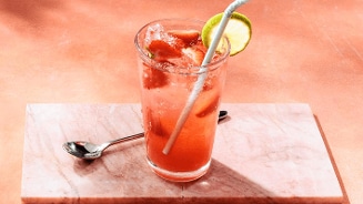 Glas met ijskoude aardbeienlimonade, stukjes aardbei, schijfje limoen en rietje op een marmeren plateau, met een lepel ernaast.