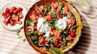 Salade met watermeloen, aardbeien, burrata, verse kruiden en granen in een grote schaal, geserveerd met een bordje aardbeien ernaast.