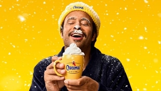 Gele mok met Chocomel-logo gevuld met slagroom, in handen van persoon met Chocomel-muts tegen gele achtergrond met sneeuwvlokken