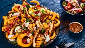 Gerecht met gekruide krulfriet, stukjes kip, granaatappelpitjes, rode ui, frisse yoghurtsaus en verse kruiden op een bord, geserveerd met een salade en kruidenmix op een blauwe tafel