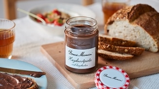 Glazen pot Bonne Maman hazelnootpasta met wit hazelnotenlabel op een ontbijttafel, naast vers gesneden brood, een bord met besmeerde boterham en glazen met drinken.