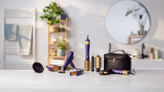 Set haartools met diverse opzetstukken en een zwarte opbergetui op een badkamerblad, met een spiegel en planten op de achtergrond