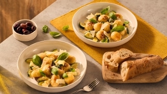 Twee borden met gnocchi, courgette, basilicum en kaas, geserveerd naast een houten plank met vers stokbrood en een schaaltje zwarte olijven op een grijs oppervlak.