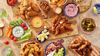 Bord met verschillende borrelhapjes zoals kipnuggets, kippenvleugels, kaasblokjes, gesneden groenten met dipsaus, plakjes worst, druiven, augurken, hummus en noten, geserveerd op een tafel voor een borrel of feestje.