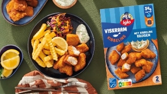 Bord met kibbeling van het merk Iglo, geserveerd met frietjes, partjes citroen, salade en een bakje saus, naast de verpakking van Iglo Viskraam Kibbeling met kibbelingkruiden.