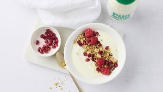 Kom met yoghurt, granola, frambozen en granaatappelpitten, naast een kleine schaal met extra granaatappelpitten, een gouden lepel en een fles melk op een lichte ondergrond.