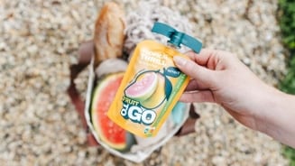 Hand houdt een zakje Fruit to Go Tropical Thrill smoothie van Servero boven een boodschappentas met stokbrood en watermeloen