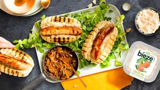 Drie gegrilde broodjes met worst, geserveerd op een bedje van sla, met een bakje gekarameliseerde uien, een schaaltje coleslaw en een verpakking Bieze coleslaw.