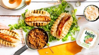 Gegrilde worstenbroodjes met gebakken uien en Bieze coleslaw op een bord, geserveerd op een wit houten tafel.