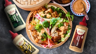 Een bamboemand gevuld met een kleurrijke salade van groenten, stukjes krokant vlees, geserveerd met verschillende sausjes van het A-merk Zaandam, waaronder Oriental, Lemon Curry en Korean BBQ. Op de achtergrond staat een schaaltje met saus en een lepel.