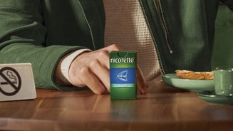 Persoon houdt een doosje Nicorette zuigtabletten vast aan een tafel, naast een bord met ontbijt en een mok