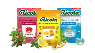 Drie verpakkingen Ricola Alpenkruiden keelpastilles in de smaken cranberry, honing citroen echinacea en fresh mint, met bijbehorende kruiden, citroen en muntblaadjes op de voorgrond.