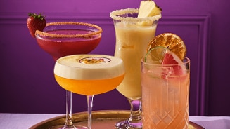 Vier kleurrijke cocktails op een gouden dienblad, elk gegarneerd met vers fruit zoals aardbei, ananas, passievrucht, limoen en sinaasappel, tegen een paarse achtergrond