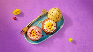 Paasdessert met vrolijke decoraties, waaronder paaseitjes en een kuikentje van marsepein, geserveerd op een blauw bord met een paarse ondergrond.