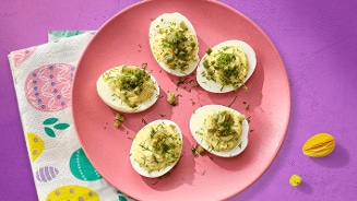 Gevulde eieren met kruiden op een roze bord, geplaatst op een paars tafelblad met een kleurrijke servet met paasdecoraties.