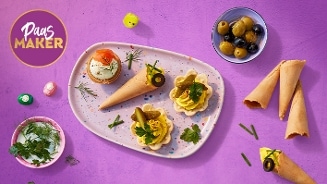 Paas hapjes op een paars oppervlak, geserveerd op een kleurrijk bord met verschillende hartige snacks, zoals gevulde hoorntjes, mini taartjes, olijven en roomkaas, versierd met verse kruiden. Links staat een klein bakje met kruiden en rechtsboven een schaaltje met groene en zwarte olijven. De sfeer is feestelijk en past bij Pasen.