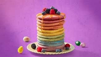 Stapel kleurrijke pannenkoeken met honing, versierd met verse blauwe bessen, frambozen en bramen op een wit bord, tegen een paarse achtergrond. Naast het bord liggen paaseitjes en een decoratief gele kuikentje.