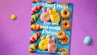 Paasfolder van Albert Heijn op een paarse ondergrond, met gekleurde croissants, bagels en paaskoekjes in de vorm van konijntjes. Tekst: "Een wel héél vrolijk Pasen". Decoratieve paasversiering en gekleurde eitjes rondom de folder.
