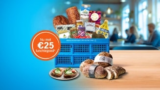 Een blauwe Albert Heijn krat gevuld met diverse lunchproducten zoals brood, zuivel, fruit en beleg, op een houten tafel met een sticker waarop staat "Nu met €25 lunchtegoed". Op de voorgrond liggen verschillende soorten brood en een bord met belegde boterhammen.