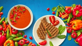 Groentesoep met tomaat en gegrilde sandwich op een bord, omringd door een verscheidenheid aan verse groenten zoals paprika's en spinazie.