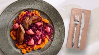 Boerenkoolstamppot met worst, rode kool en stukjes appel op een grijs bord, met bestek en een servet ernaast