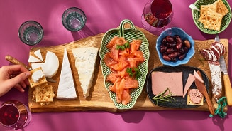 Borrelplank met verschillende kazen, crackers, gerookte zalm, olijven, paté, worst en glazen rode wijn op een roze ondergrond