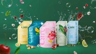 Vijf kleurrijke Care handzeep navulverpakkingen met varianten Lemon, Bergamot Boost, Cherry Blossom, Botanical Sage en Mint, omringd door verse ingrediënten zoals citroen, gember, munt, kersen, kruiden en bloemblaadjes op een groene achtergrond.