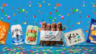 Mensen verkleed als worstenbroodjes tussen verschillende Albert Heijn producten zoals een pizza, Brouwers 0.0 bier, AH runder knakworsten, AH scharreleieren en AH Chips chips, met confetti op een blauwe achtergrond.