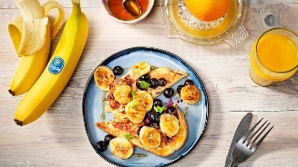 Ontbijtgerecht van pannenkoeken met plakjes gebakken banaan en blauwe bessen, naast Chiquita-bananen, een glas vers geperste sinaasappelsap en een kommetje honing.